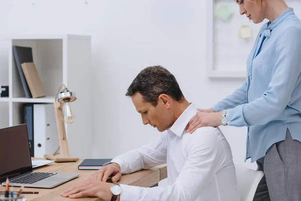 Hombre de negocios que trabaja con el ordenador portátil en el lugar de trabajo, mientras que su colega haciendo masaje para él - foto de stock