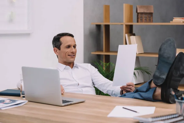 Красивый улыбающийся бизнесмен читает документ на рабочем месте в офисе — стоковое фото