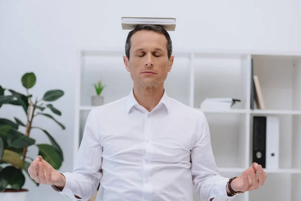 Handsome businessman meditating with book on head at office — Stock Photo