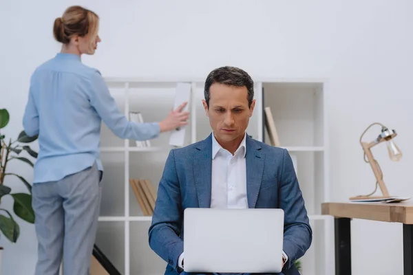 Homem de negócios trabalhando com laptop, enquanto seu colega à procura de documentos em estantes no escritório moderno — Fotografia de Stock
