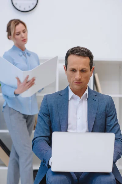 Бізнесмен працює з ноутбуком, коли його колега читає документи в сучасному офісі — стокове фото