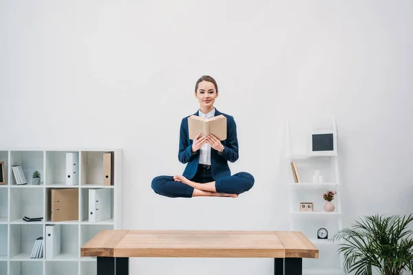 Lächelnde junge Geschäftsfrau liest Buch, während sie am Arbeitsplatz schwebt — Stockfoto