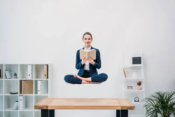 Souriant jeune femme d'affaires lecture livre tout en lévitant sur le lieu de travail — Photo de stock