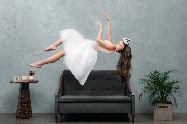 Beautiful  barefoot girl in virtual reality headset levitating above couch — Stock Photo