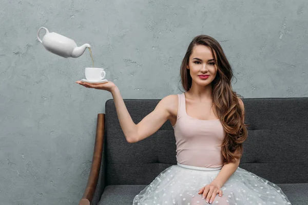Beautiful woman smiling at camera and holding cup while levitating teapot pouring tea — Stock Photo