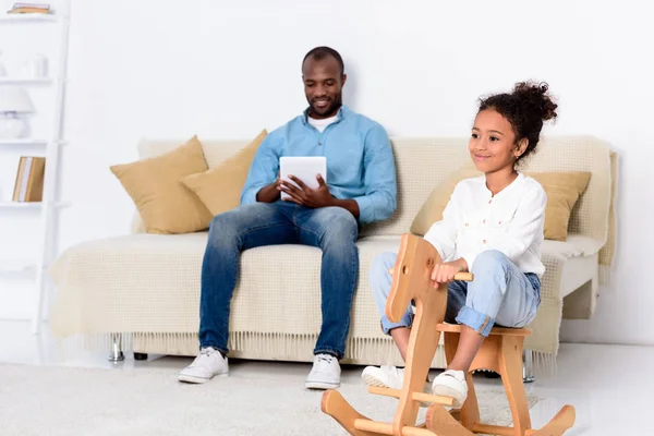 African american father using tablet and daughter sitting on rocking horse — Stock Photo