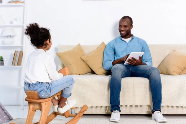 Afrikanisch-amerikanischer Vater mit Tablet sieht Tochter zu Hause auf Schaukelpferd sitzen — Stockfoto