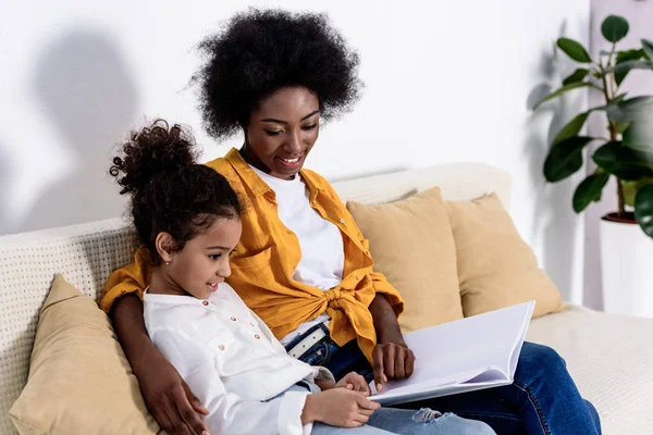 Afro-americana mãe e filha lendo livro no sofá em casa — Fotografia de Stock