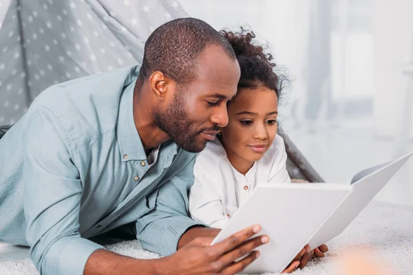 African american father and daughter lying on floor and reading together at home — Stock Photo