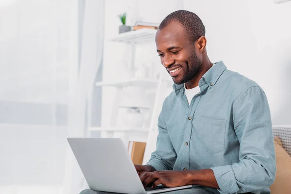 Hombre afroamericano feliz usando el ordenador portátil en el sofá en casa - foto de stock