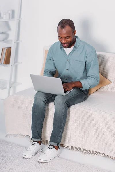 Heureux homme afro-américain en utilisant un ordinateur portable sur le canapé à la maison — Photo de stock