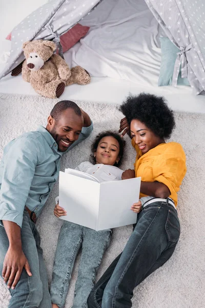 Visão de alto ângulo dos pais e filha afro-americanos deitados no chão e livro de leitura — Fotografia de Stock
