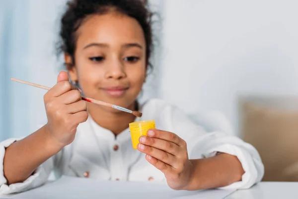 Adorable african american kid putting paint brush into yellow poster paint at home — Stock Photo