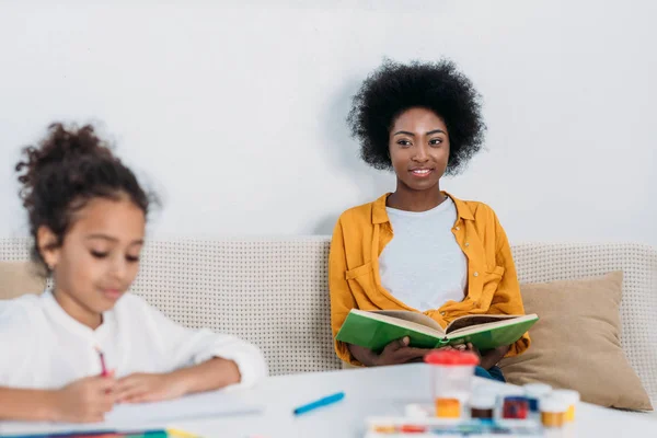 Madre afroamericana leyendo libro y dibujo de hija con plumas de fieltro en casa - foto de stock