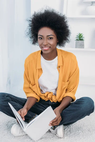 Lächelnde Afroamerikanerin mit Buch in der Hand und Blick in die Kamera zu Hause — Stockfoto