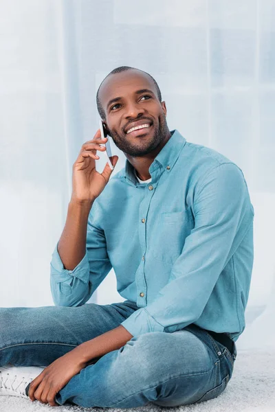 Homme afro-américain souriant assis sur le sol et parlant par smartphone à la maison — Photo de stock