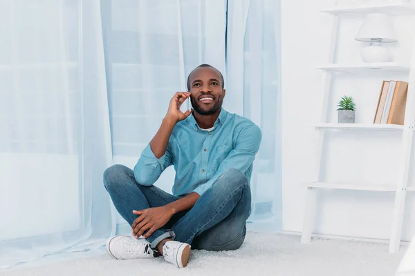 Улыбающийся африканский американец сидит на полу и разговаривает по смартфону дома — стоковое фото