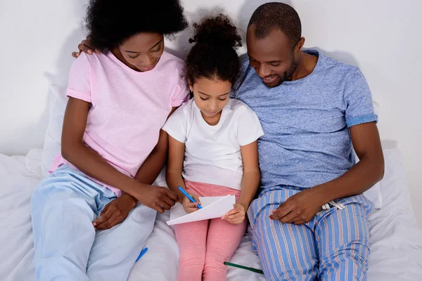 Afro-americanos pais ajudando filha pintura no álbum na cama em casa — Fotografia de Stock