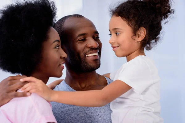 Padres afroamericanos y su hija abrazándose y mirándose en casa - foto de stock