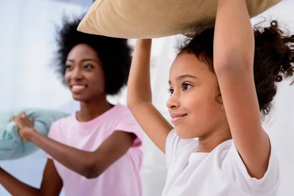African american mother and daughter holding pillows and looking away at home — Stock Photo