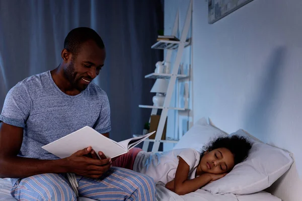 African american father reading book for sleeping daughter at home — Stock Photo