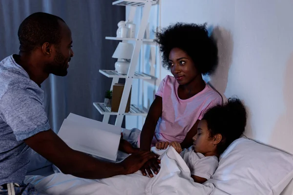 Padres afroamericanos con libro e hija en la cama - foto de stock