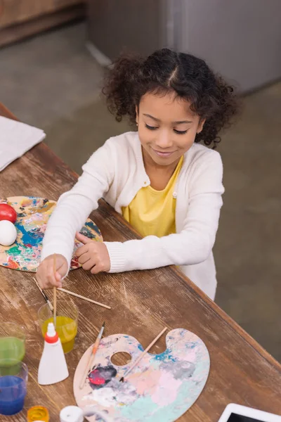 High angle view of african american kid putting brush into glass with paint for painting easter eggs — Stock Photo