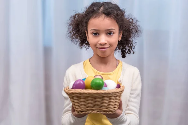 Adorable african american kid holding straw basket with painted easter eggs and looking at camera — Stock Photo