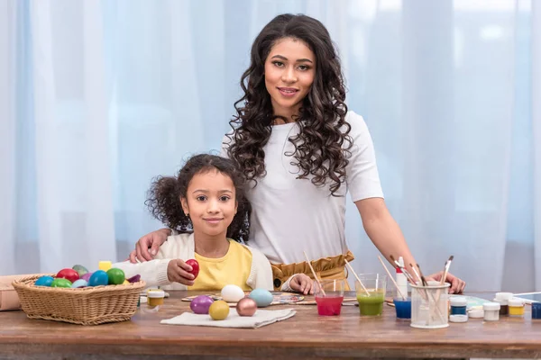 Afro-americana mãe e filha de pé perto da mesa com ovos de Páscoa e olhando para a câmera — Fotografia de Stock