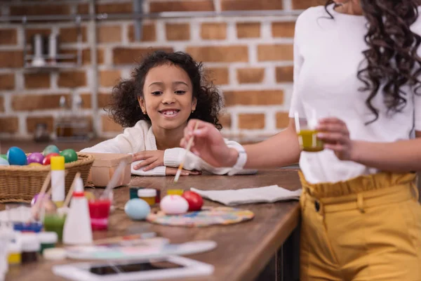 Обрезанное изображение африканской американской дочери, смотрящей, как мама рисует пасхальные яйца — стоковое фото