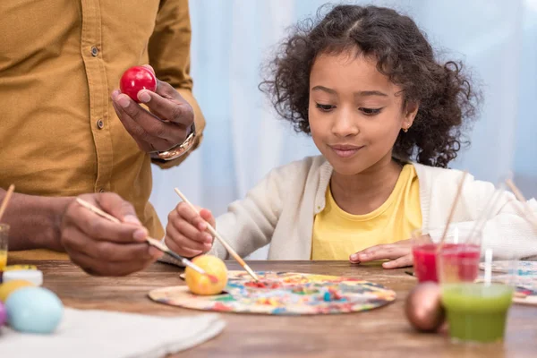 Image recadrée du père afro-américain et adorable fille peignant des œufs de Pâques avec des peintures par affiches — Photo de stock