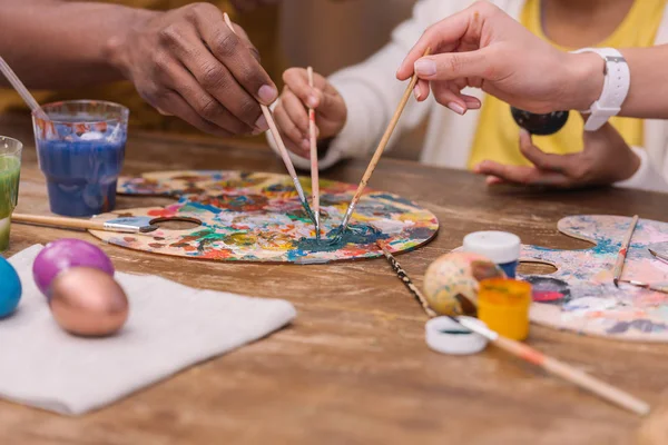 Cropped image of african american parents and daughter putting paint brushes on palette, easter concept — Stock Photo