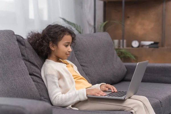 Vista lateral del niño afroamericano usando el ordenador portátil en el sofá en casa - foto de stock