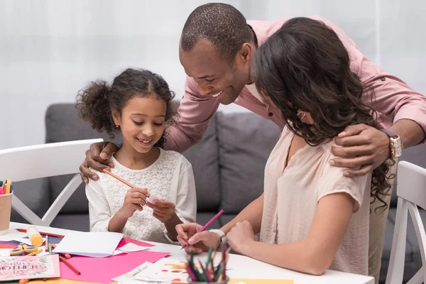 Afroamericanos padres mirando cómo hija dibujo parte para tarjeta de felicitación - foto de stock