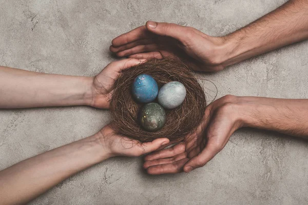 Vista recortada de pareja sosteniendo nido con huevos de Pascua pintados en superficie de hormigón - foto de stock