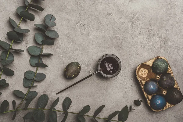 Top view of painted easter eggs in golden tray with eucalyptus, paint and brush on concrete — Stock Photo