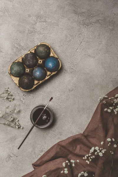 Vista superior de ovos de páscoa pintados em bandeja com tinta, pincel e flores em pano — Fotografia de Stock