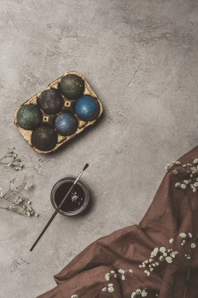 Top view of painted easter eggs in tray with paint, brush and flowers on cloth — Stock Photo