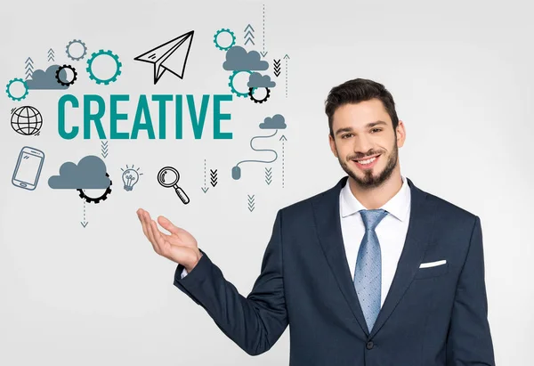 Handsome young businessman showing creative icons and smiling at camera isolated on grey — Stock Photo