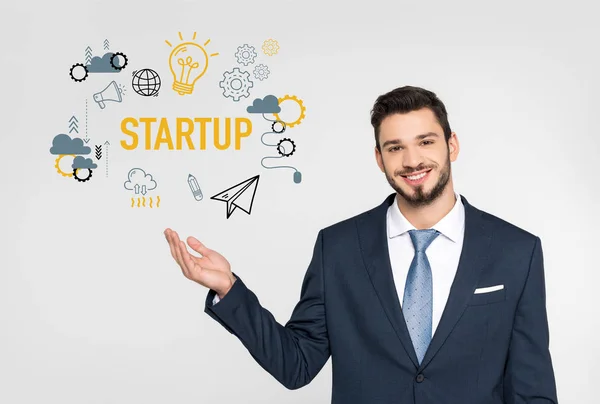 Handsome young businessman showing startup icons and smiling at camera isolated on grey — Stock Photo