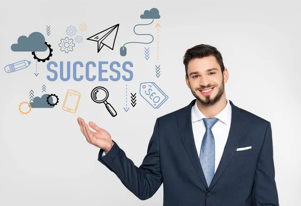 Handsome young businessman showing success icons and smiling at camera isolated on grey — Stock Photo