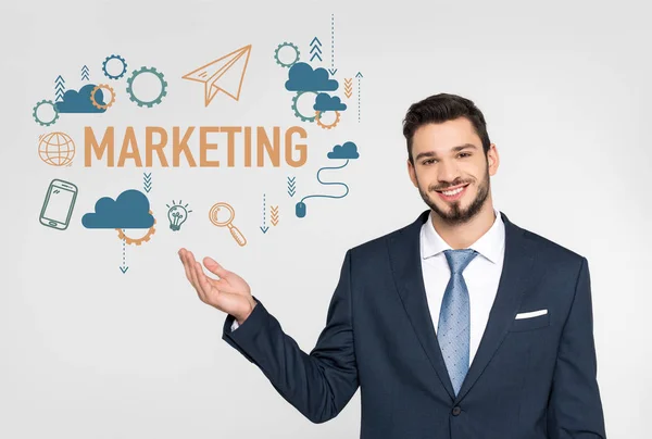 Handsome young businessman showing marketing icons and smiling at camera isolated on grey — Stock Photo