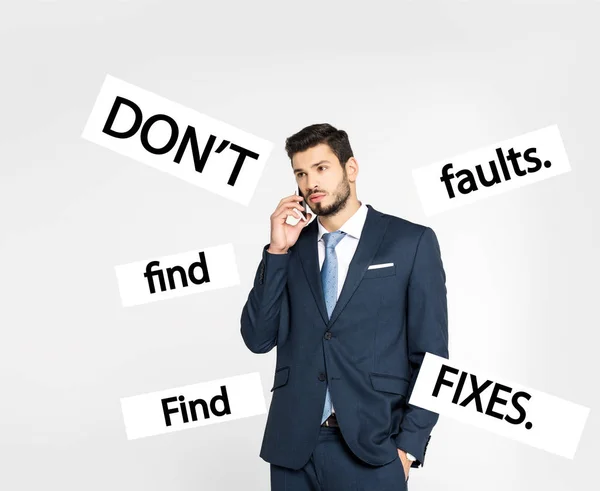 Stylish young businessman standing with hand in pocket and talking on smartphone while looking away isolated on grey, dont find faults. find fixes inscription — Stock Photo