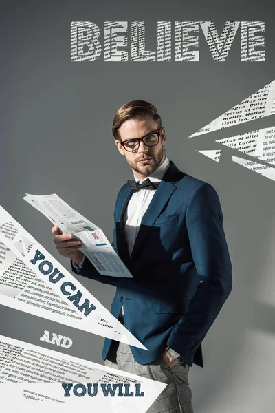 Portrait of handsome fashionable businessman in spectacles reading newspaper and motivational inscription on grey — Stock Photo