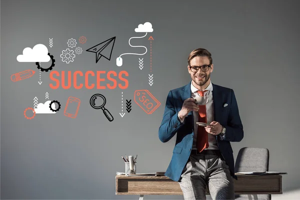 Smiling businessman with cup of coffee sitting on table and looking at camera, success icons on grey — Stock Photo