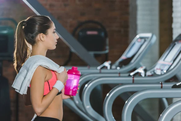 Sporty woman with towel and bottle of water standing against treadmill — Stock Photo