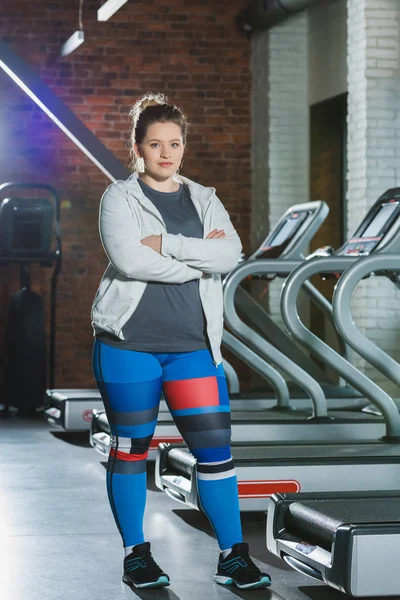 Overweight woman standing against treadmills with arms crossed — Stock Photo