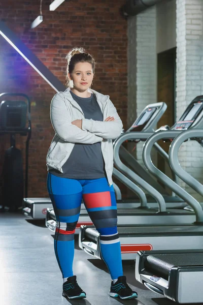 Femme en surpoids debout contre des tapis de course avec les bras croisés — Photo de stock