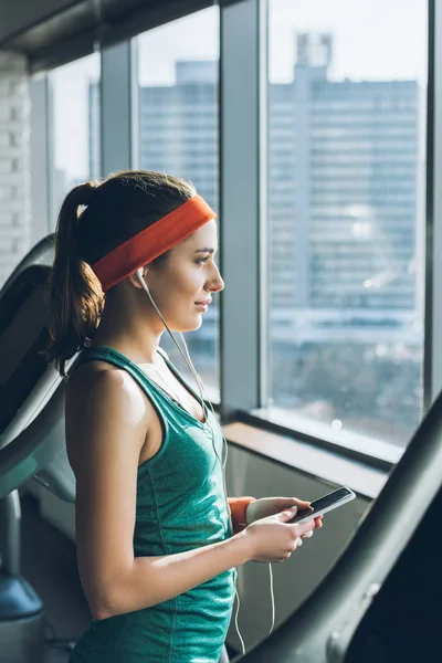 Donna sportiva che utilizza smartphone e auricolari in palestra mentre guarda la finestra — Foto stock