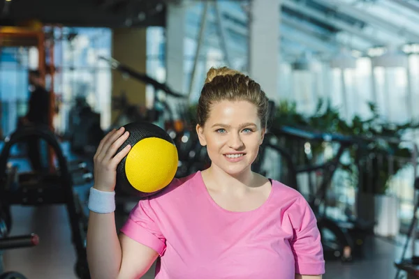 Sonriente mujer con sobrepeso con balón de medicina en el hombro - foto de stock